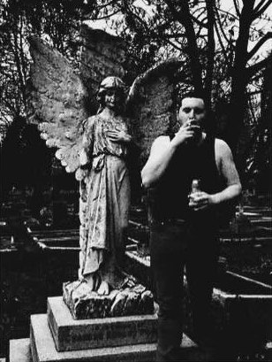 Promo photo: figure smoking beside an angel statue in a graveyard